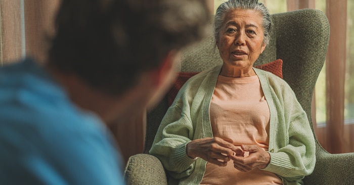 An elderly female nursing home resident sits in a green armchair and talks facing a female professional caregiver. 