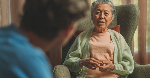 An elderly female nursing home resident sits in a green armchair and talks facing a female professional caregiver. 