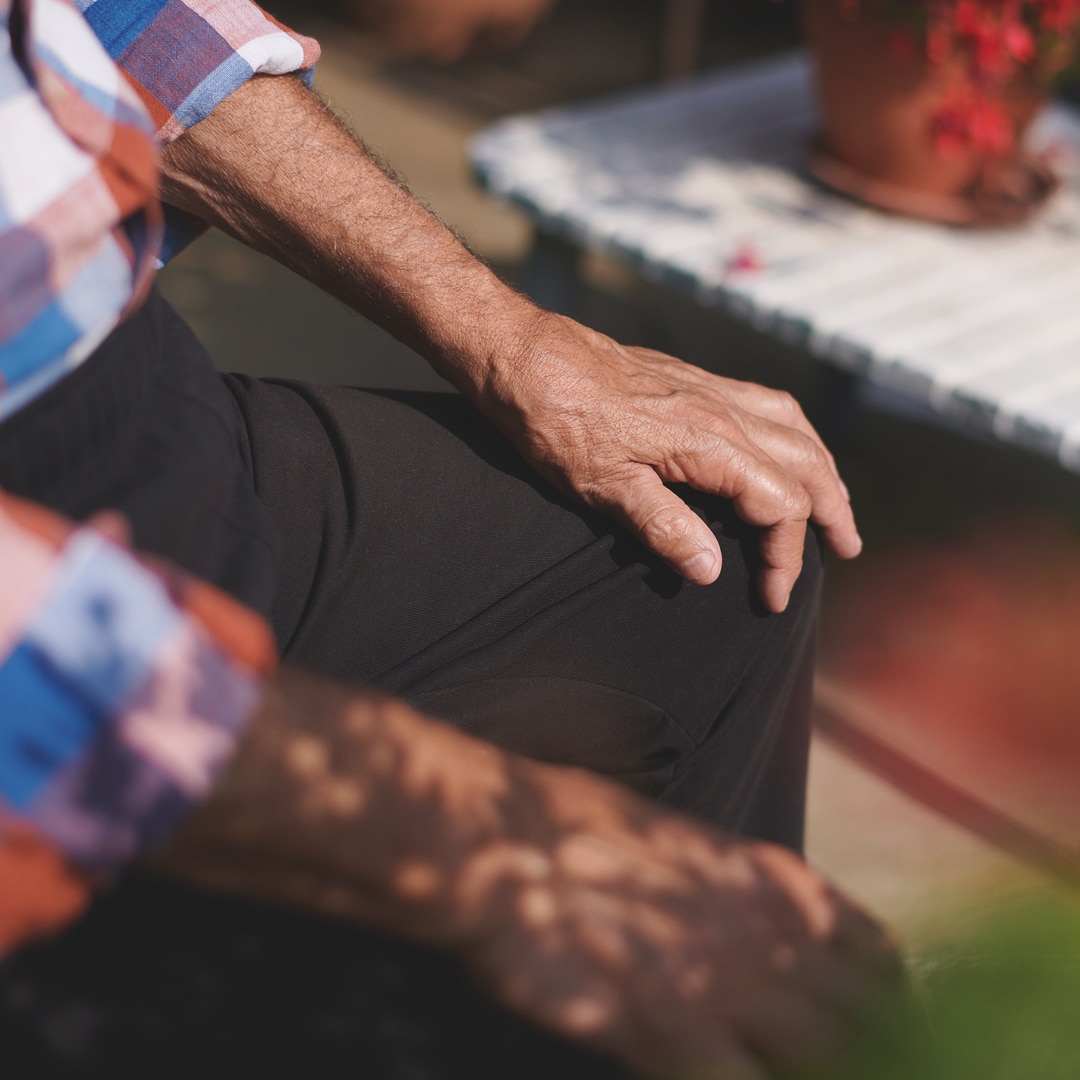 A close-up of an elderly person's hand resting on their knee. The hand appears slightly wrinkled, and the person is wearing a plaid shirt and dark pants. Sunlight filters through leaves, casting dappled shadows on the arm. A white outdoor table with a potted plant is visible in the softly blurred background.