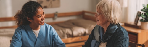 A female professional caregiver chats with an elderly female nursing home resident in the resident’s room