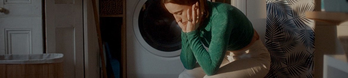 Pavargusi moteris sėdi viena skalbimo kambaryje.