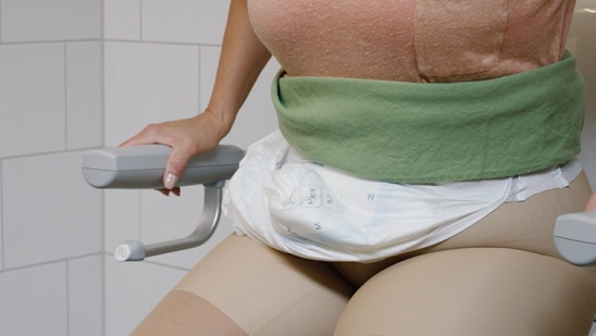 A close-up of a person sitting on a toilet with assistive armrests, wearing a disposable adult diaper and compression stockings. Their hands grip the armrests for support. The background features white tiled walls, indicating a bathroom setting.