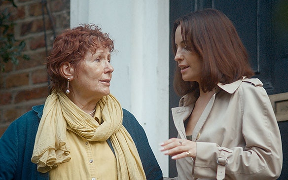 An older woman in a yellow scarf talks with a younger woman in a beige trench coat outside a building.
