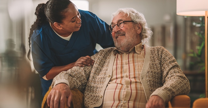 A female professional caregiver wearing a blue uniform leans over the shoulder of a male nursing home resident sitting in an armchair. They talk and laugh together. 