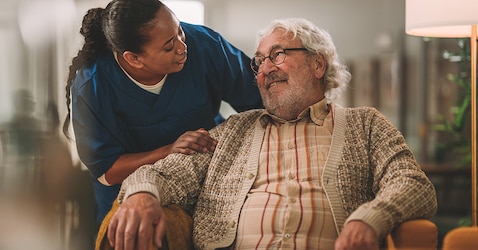 A female professional caregiver wearing a blue uniform leans over the shoulder of a male nursing home resident sitting in an armchair. They talk and laugh together. 