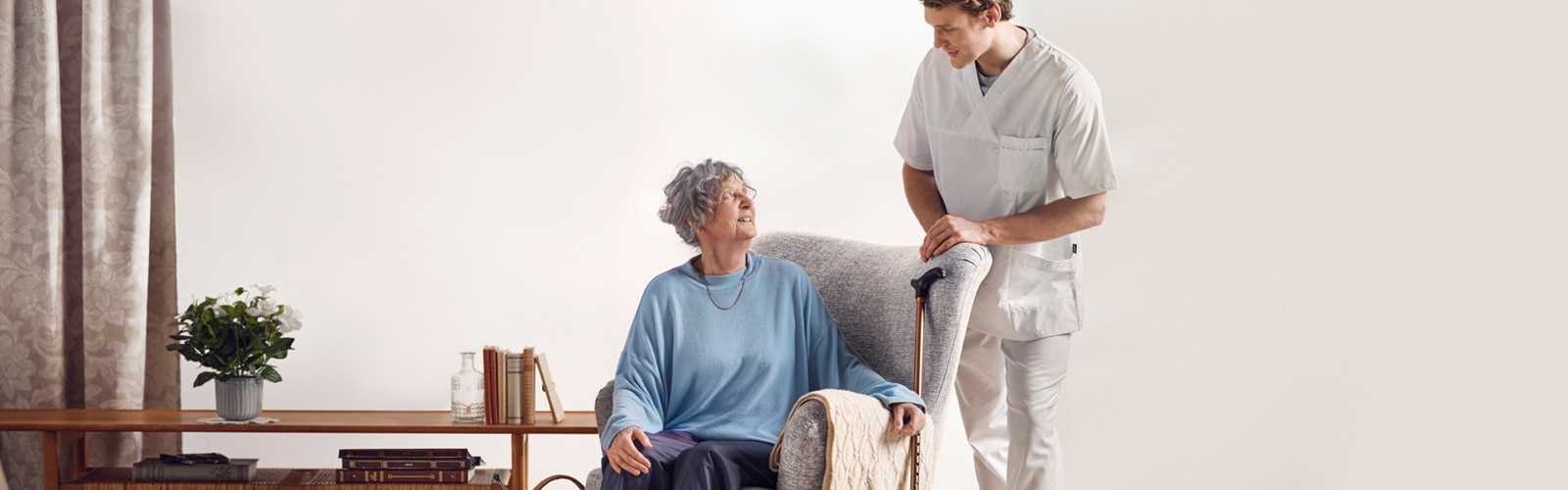 A seated elderly resident talking with a standing professional caregiver in a care home environment 
