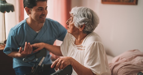 A smiling male professional caregiver supports and holds the hand of a female nursing home resident sitting in her bed. 