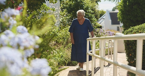An elderly female nursing home resident walks in the nursing home garden. It is a sunny day and there are plants and flowers. 