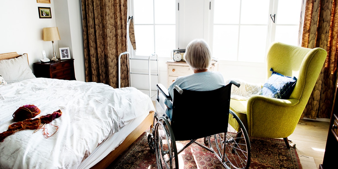 Seniorin im Rollstuhl sitzt in einem freundlich, hellen Zimmer und schaut in Richtung Fenster