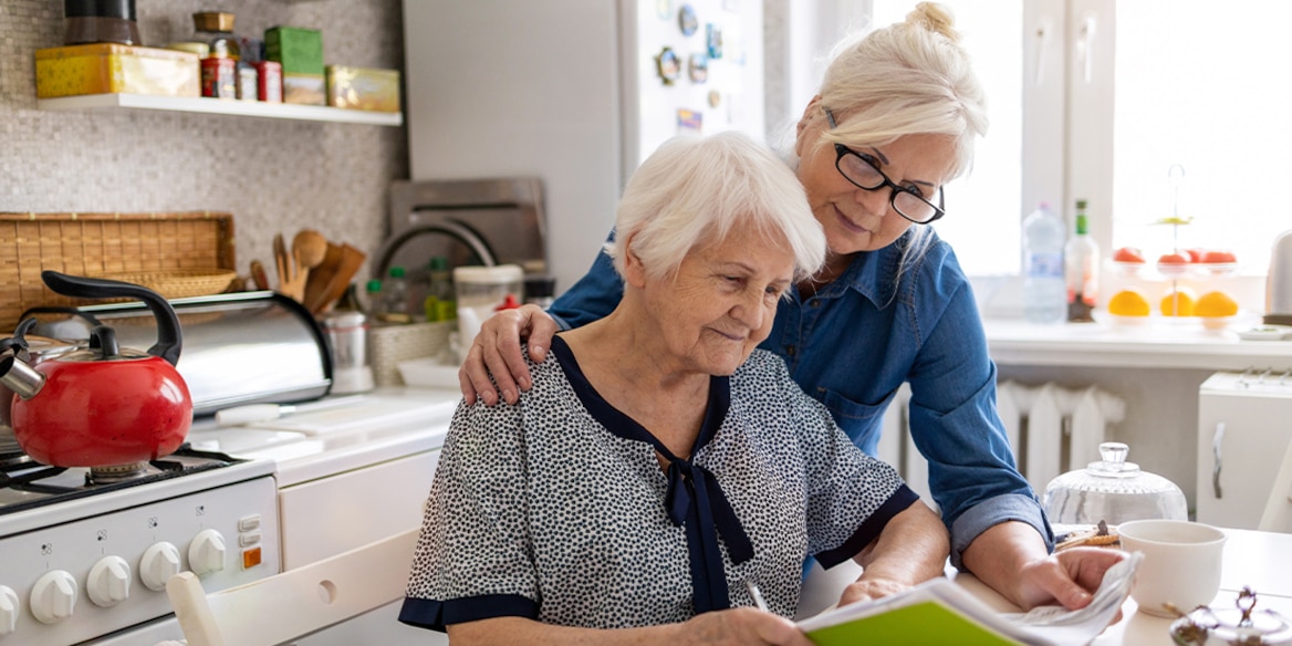 Eine Frau mit Brille schaut mit ihrer Mutter am Küchentisch in Unterlagen und legt ihre Hand auf die Schulter