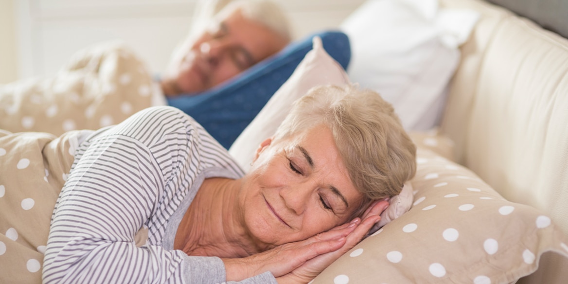 Ein Seniorenpaar schläft friedlich im Bett – sie vorn, er im Hintergrund leicht unscharf.