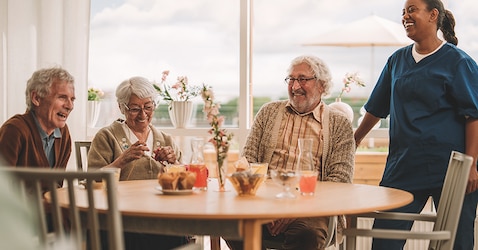 A group of male and female nursing home residents sit around a dining table. They smile and laugh while a female professional caregiver stands smiling. 