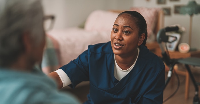 A smiling female professional caregiver kneels down in front of an elderly female nursing home resident. She talks in a reassuring manner. 