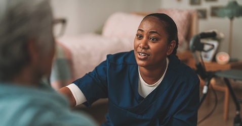 A smiling female professional caregiver kneels down in front of an elderly female nursing home resident. She talks in a reassuring manner. 