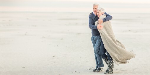 Un homme âgé prend sa femme dans ses bras pour la réchauffer tandis qu’ils marchent sur une plage balayée par le vent