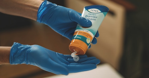 An image showing a professional caregiver’s hands. They are wearing protective gloves and are squeezing barrier cream from a tube. 