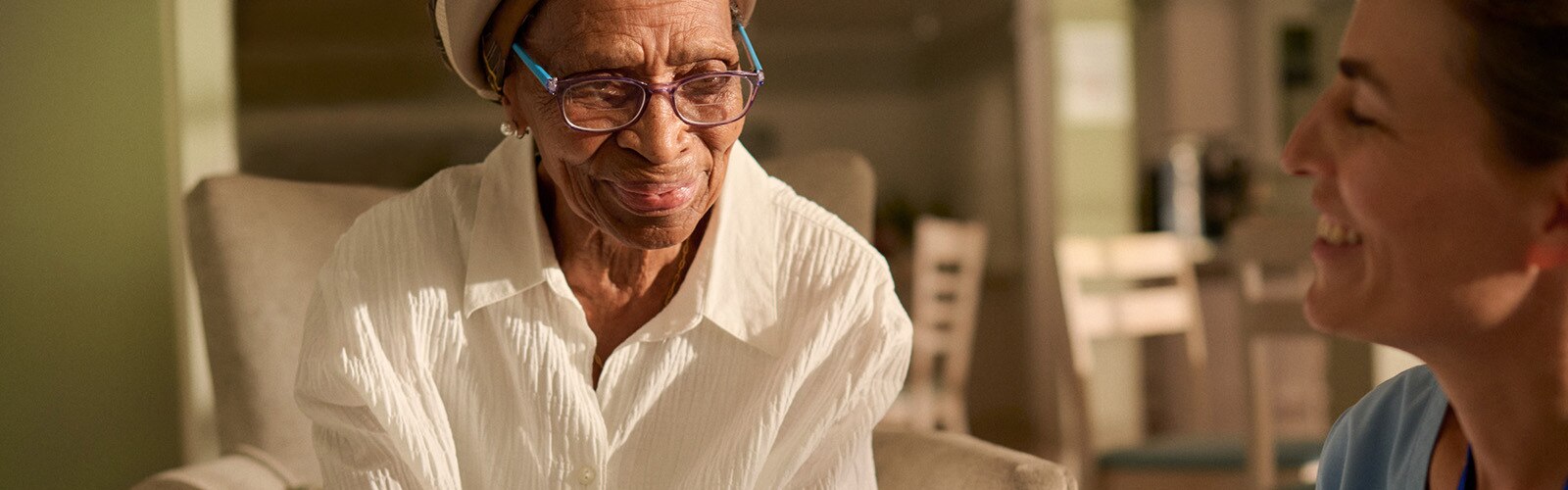 En äldre kvinna med glasögon och vit skjorta sitter i en fåtölj och ler varmt under ett samtal med en vårdpersonal i blå arbetskläder i en omsorgsmiljö.