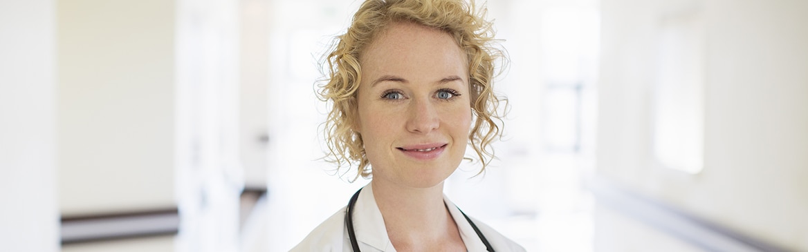 Eine blonde Ärztin mit Stethoskop steht auf einem Krankenhausflur und lächelt