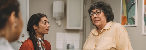 An elderly woman sits on an exam table in a medical office, looking concerned. A younger woman in a red sweater listens attentively, while a nurse is slightly out of focus in the foreground. The setting includes medical equipment, indicating a clinical environment.