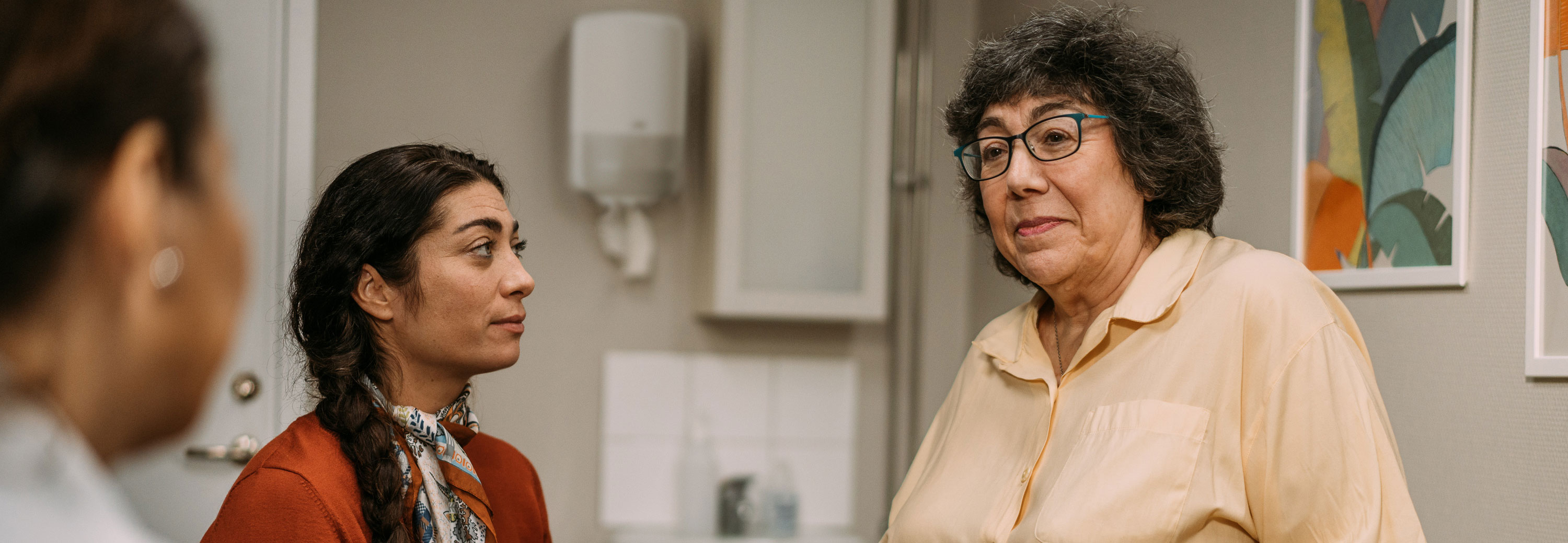 An elderly woman sits on an exam table in a medical office, looking concerned. A younger woman in a red sweater listens attentively, while a nurse is slightly out of focus in the foreground. The setting includes medical equipment, indicating a clinical environment.