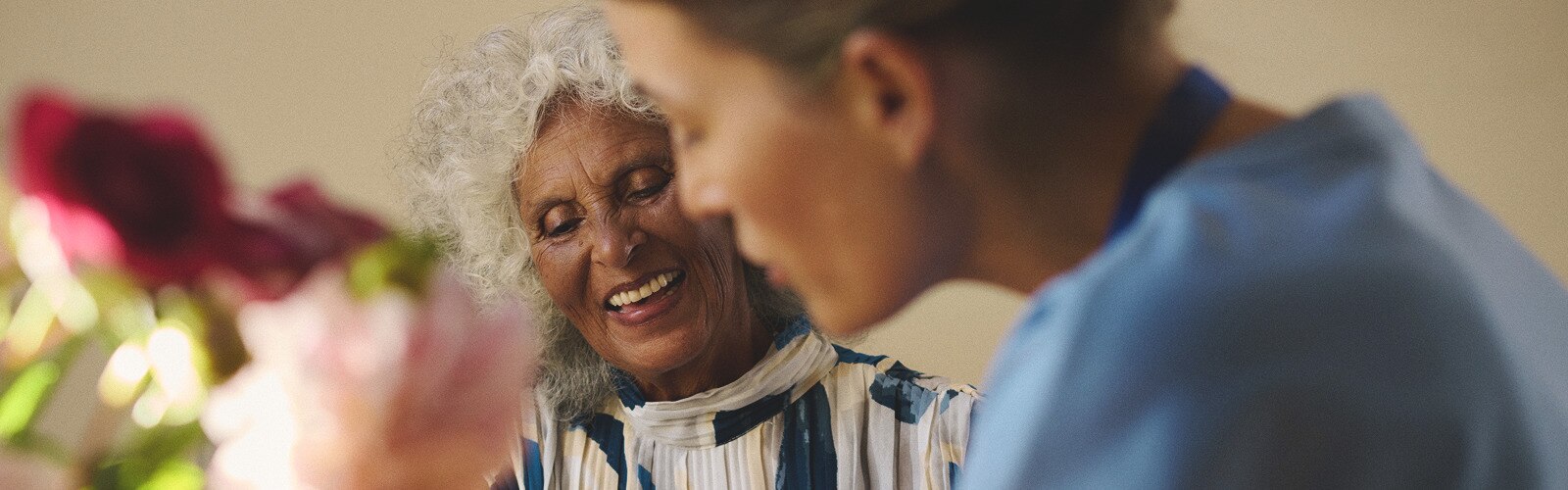 A female professional caregiver and an elderly female nursing home resident sit at a table with flowers while they talk together and smile. 