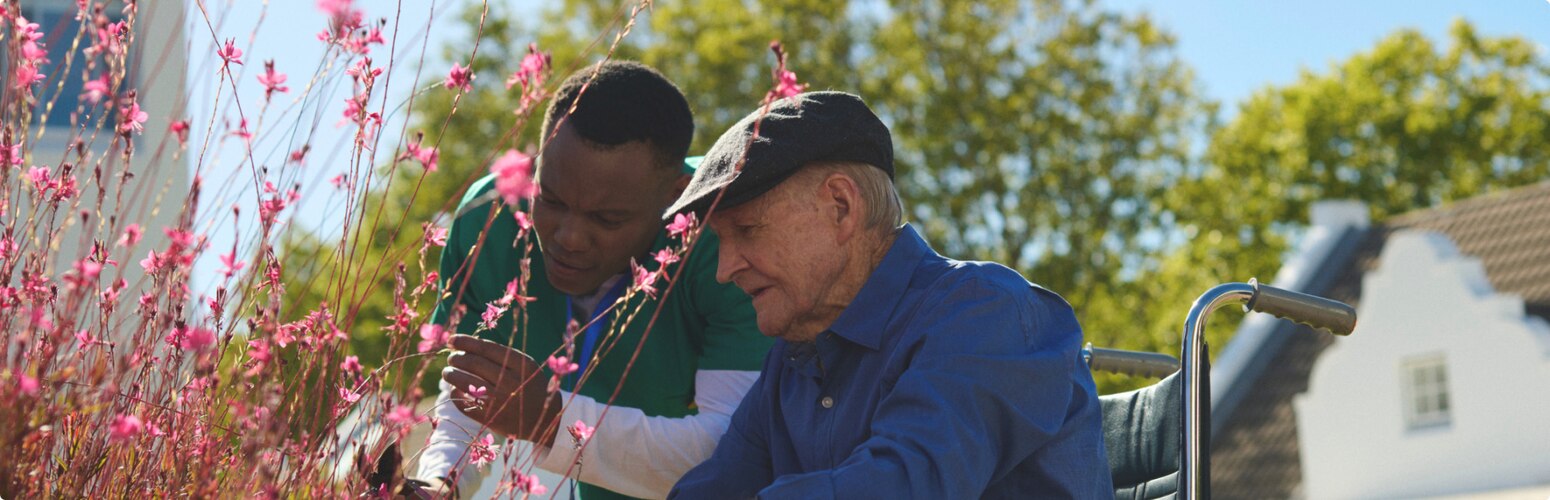 A professional caregiver and an elderly male nursing home resident in a wheelchair look at flowers together in a garden