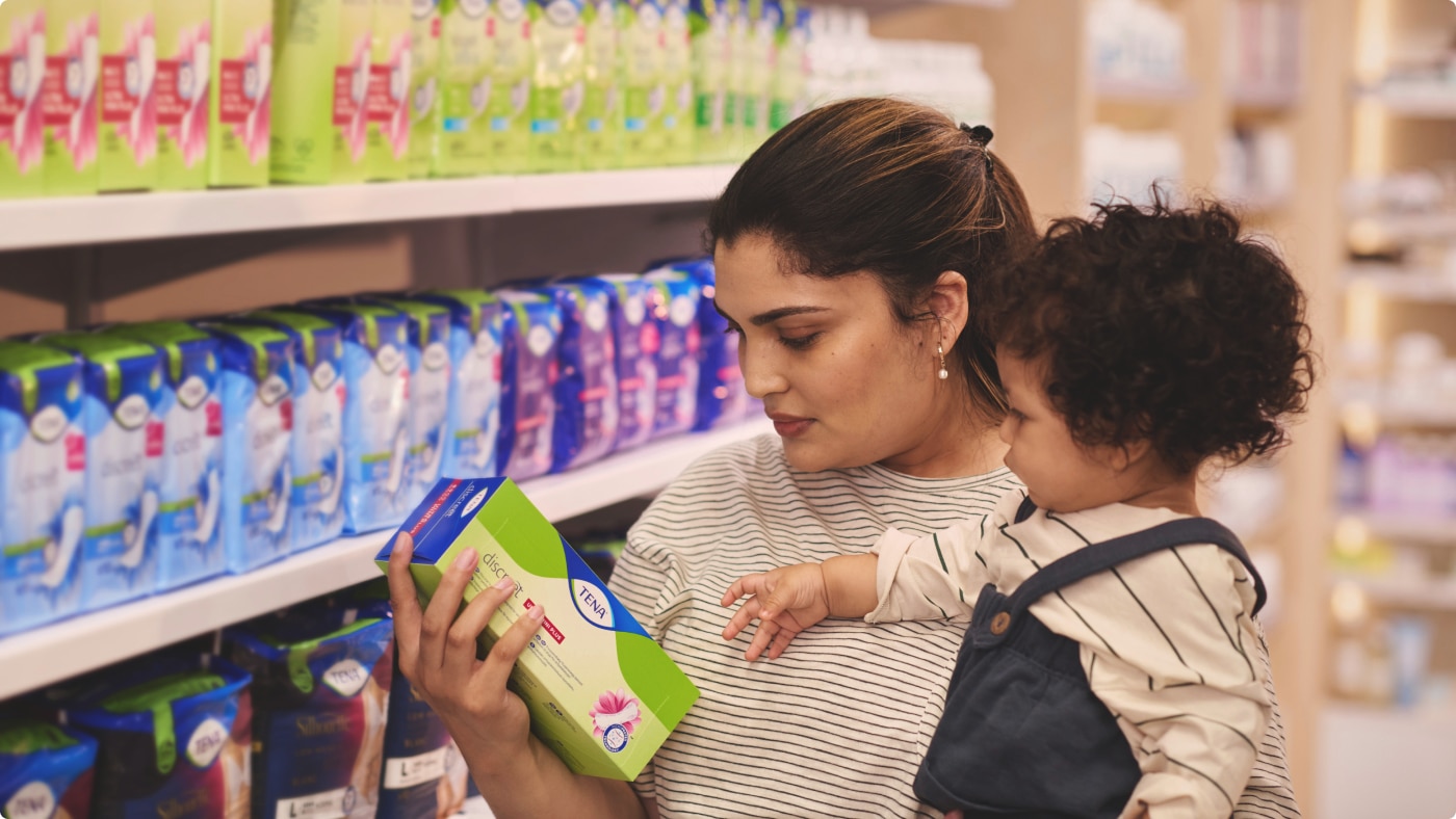 Une femme tient son bébé dans une pharmacie tout en regardant les protections contre l’incontinence TENA Discreet.