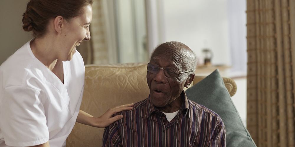 Caregiver in a white uniform laughing with an elderly man sitting on a couch, both appearing joyful and engaged.