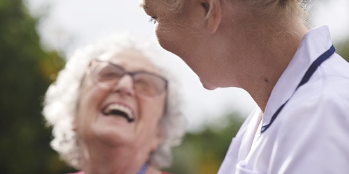 En äldre kvinnlig vårdtagare och hennes sjuksköterska skrattar utomhus.