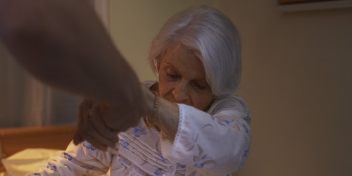 An older woman is assisted out of her bed by a caregiver