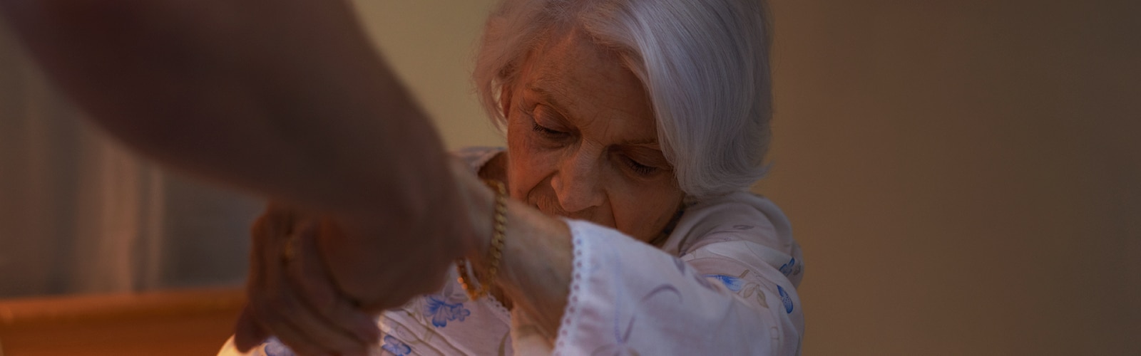 An elderly woman with white hair sitting on a bed, wearing a floral-patterned nightgown. She is being assisted by another person, whose hand is extended to help her.