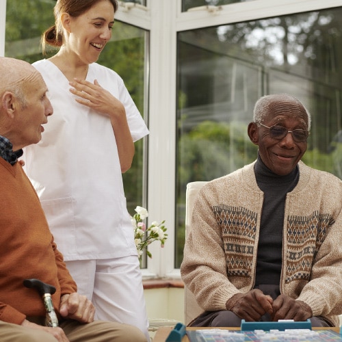 A healthcare worker in white scrubs stands between two elderly men in a sunlit room with large windows. The men are seated, one wearing an orange top and the other in a patterned beige sweater over a black turtleneck. They appear to be engaged in a cheerful conversation, with all three people smiling. A game or activity appears to be on the table in front of them.