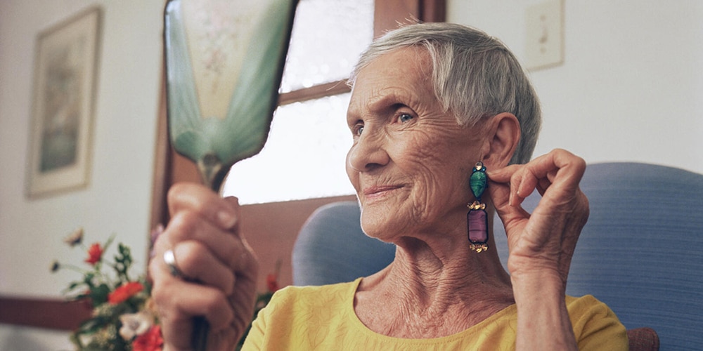 Usmívající se starší klientka si pomocí příručního zrcátka zkouší náušnice v prostředí domova pro seniory 