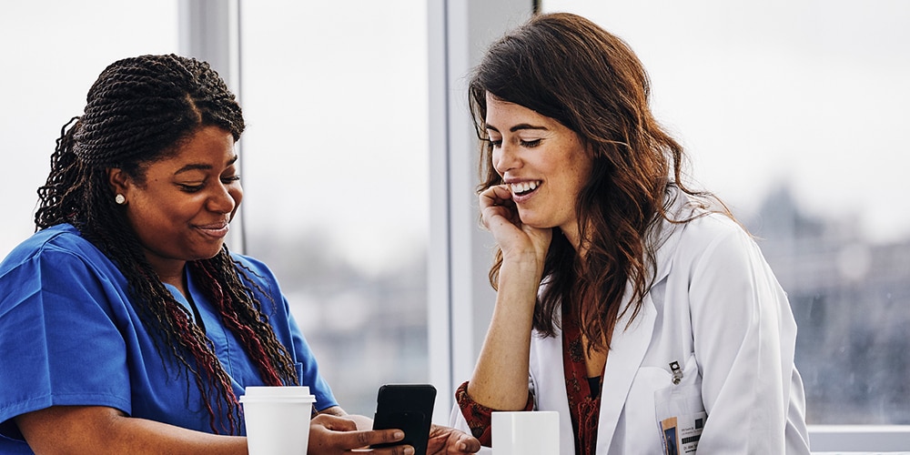 Naispuolinen terveydenhuollon ammattilainen ja naispuolinen ylihoitaja katsomassa matkapuhelinta ja keskustelemassa kanttiinissa kahvilla ollessaan 