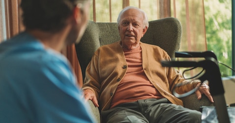 A contented, elderly male nursing home resident sits in an armchair and converses with a professional caregiver