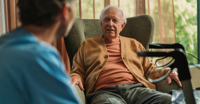 A contented, elderly male nursing home resident sits in an armchair and converses with a professional caregiver