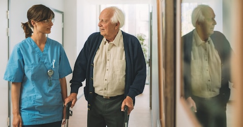 An elderly male nursing home resident uses a walking frame to walk down a corridor. He is accompanied by a female professional caregiver.