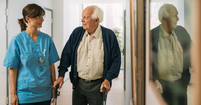 An elderly male nursing home resident uses a walking frame to walk down a corridor. He is accompanied by a female professional caregiver.