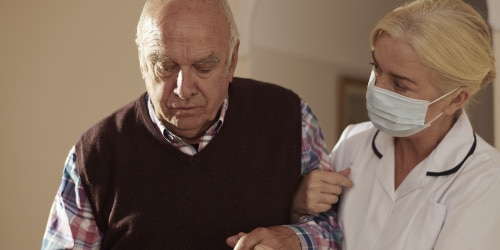 A female professional caregiver links arms and converses with an elderly male resident in a care home environment