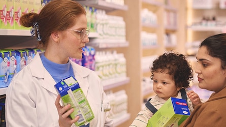 A female pharmacist in a white coat gives incontinence advice to a mother holding a young child. Both women are holding TENA products.