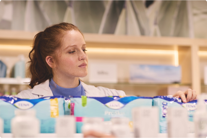 Eine Apothekerin in einer Apotheke betrachtet ein Regal, das mit TENA-Inkontinenzprodukten bestückt ist.