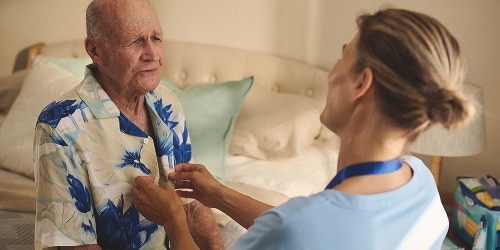 A female professional caregiver helps an elderly male nursing home resident button his shirt. They are in the resident’s room and talking and smiling together. 