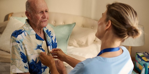 A female professional caregiver helps an elderly male nursing home resident button his shirt. They are in the resident’s room and talking and smiling together. 