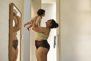 A mother with a curvy body and dark hair tied up in a bun is standing in a hallway, wearing a light green bralette and black underwear with lace details. She is lifting her baby, who has curly hair and is dressed in a beige onesie. They are looking at each other with joy.